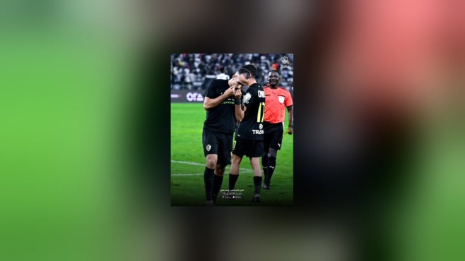 لاعب بقميص الزمالك الأسود يقبل شعار النادي، وبجواره لاعب آخر وحكم يرتدي قميصاً أحمر داخل الملعب، مع شعار الزمالك أعلى الصورة.