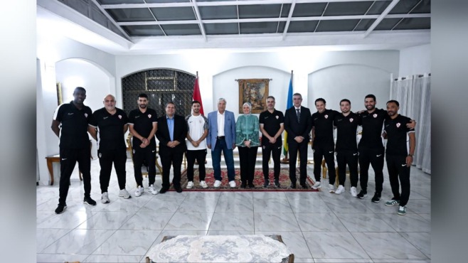 صورة جماعية داخل قاعة تضم مجموعة من لاعبي الزمالك بملابس رياضية سوداء ورجالاً بملابس رسمية وسيدة ترتدي زياً أخضر، مع علمين في الخلفية.
