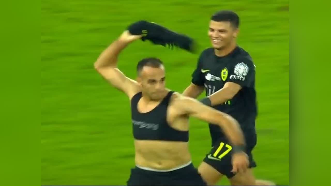 لاعب يرتدي سترة تدريب سوداء يرفع قطعة ملابس سوداء على أرضية ملعب عشبي، بينما يقف زميله خلفه ممسكاً بذراعه.
